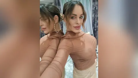 Beautiful woman in a brown long-sleeve sheer top and hoop earrings posing for a glamorous mirror selfie.