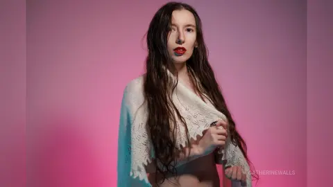 Young woman with long wavy hair wearing a white knit shawl against a soft pink studio background.