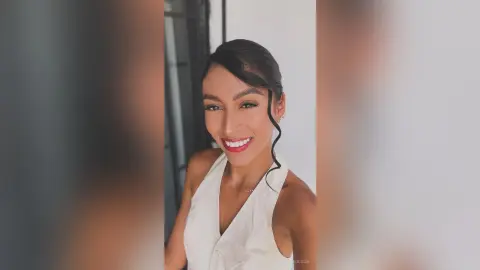 LaraOlson smiles at the camera wearing a white sleeveless top with her dark hair styled back in a wavy part.