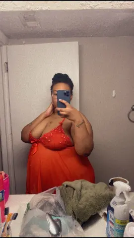 MilfArie22 poses in a red slip dress, showing cleavage while holding her phone for a mirror selfie in a bathroom setting.