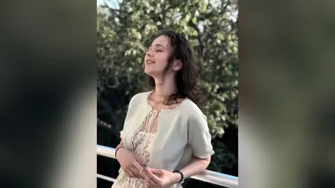 Smiling young woman in a white floral dress and light green cardigan enjoying sunny weather in a lush garden.