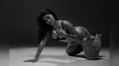 Moody black and white portrait of a woman in a black bra and denim jeans posing on a studio floor.