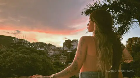 Woman in a beige tank top gazing at a sunset cityscape from a balcony with palm trees.