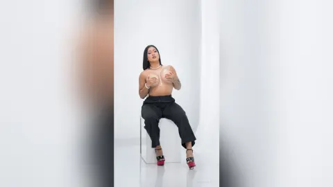 KimRoberth sits on a clear chair, holding her bare breasts while wearing black trousers and red heels.