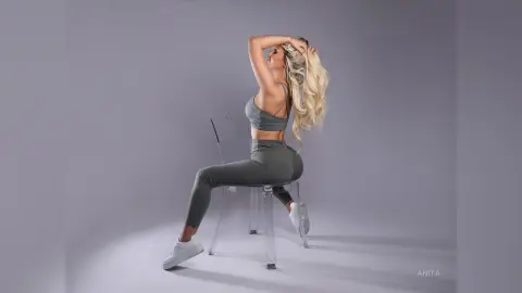 Anita sits on a clear chair wearing a grey sports bra and matching leggings, running her hands through her long blonde hair.