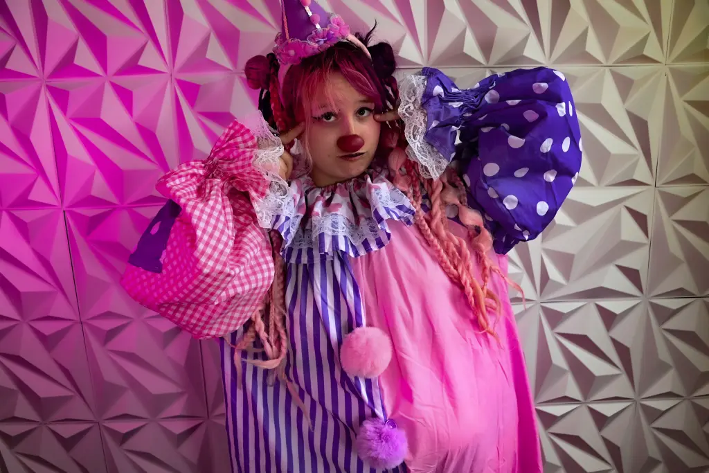 Smiling woman with pink and blue hair in gothic lace outfit posing against a colorful studio background.