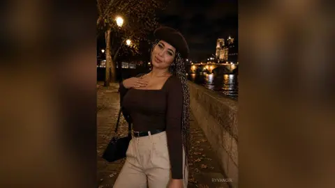 Smiling woman in a brown long-sleeve top and beige trousers posing by a river at night in Paris.