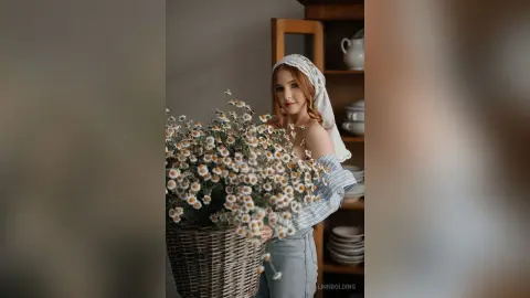 Redheaded woman in a white linen top holding a large basket of daisies in a cozy, sunlit indoor setting.
