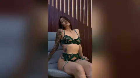 EmiliaRhodes poses in green lace lingerie and stockings while sitting on a grey sofa.