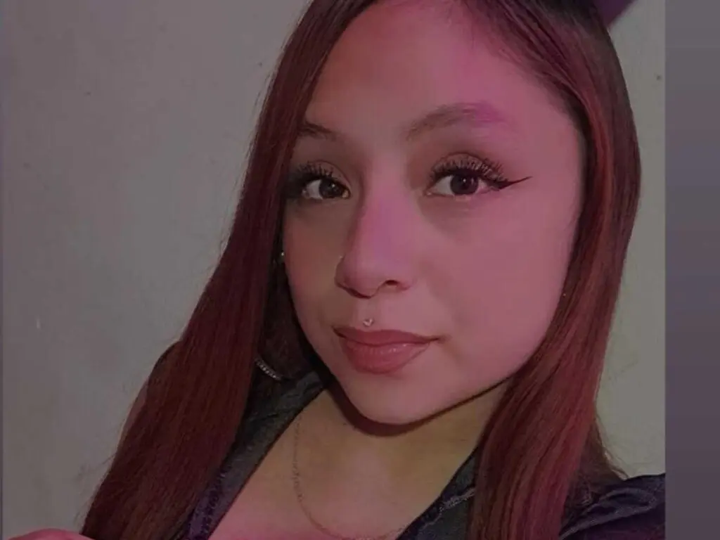 Young woman with long reddish-brown hair and winged eyeliner posing in a dark indoor setting.