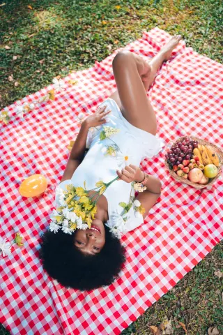 Smiling Black woman in a white dress lying on a red checkered picnic blanket with flowers and fruit in a sunny park.