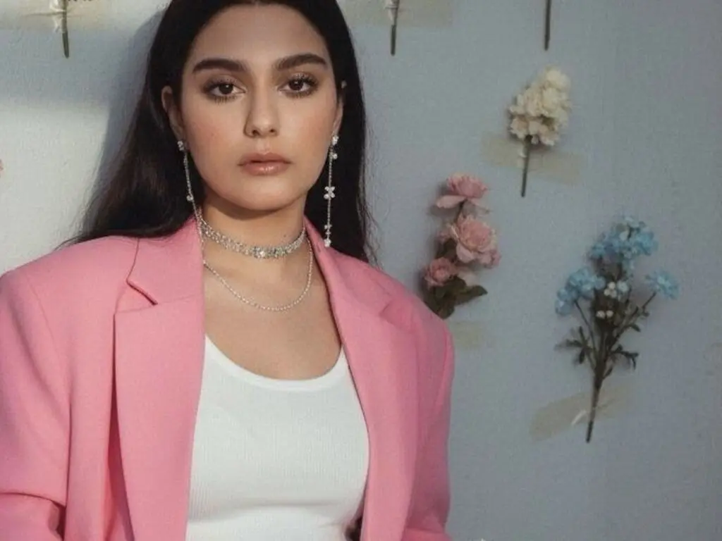 Young woman in a pink blazer and white top posing elegantly against a floral wall background.