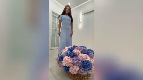 Elegant woman in a light blue midi dress standing next to a large basket of colorful hydrangeas in a bright room.