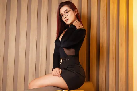 Stylish woman with red hair in a black long-sleeve top and skirt posing against a warm wooden slat wall.