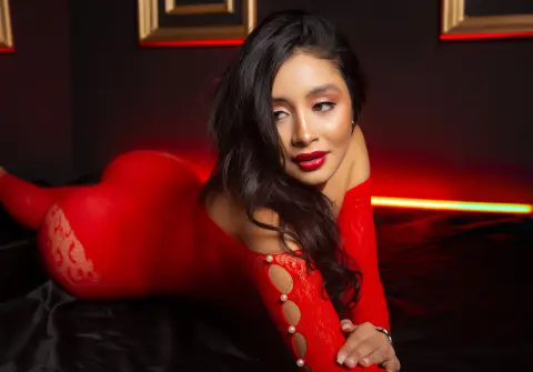 Glamorous woman in a red bodysuit lounging on black satin fabric under moody red lighting.