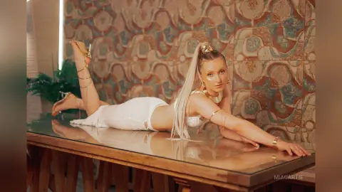 Elegant woman in a white outfit reclining on a glass table against a patterned bohemian wall background.