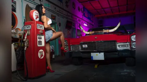 MissyJolie poses next to a red gas pump in denim shorts and a white bra, leaning one leg up against a vintage red car.