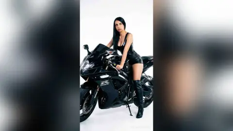 Stylish woman in black leather outfit posing confidently with a sleek black sport motorcycle on a white background.