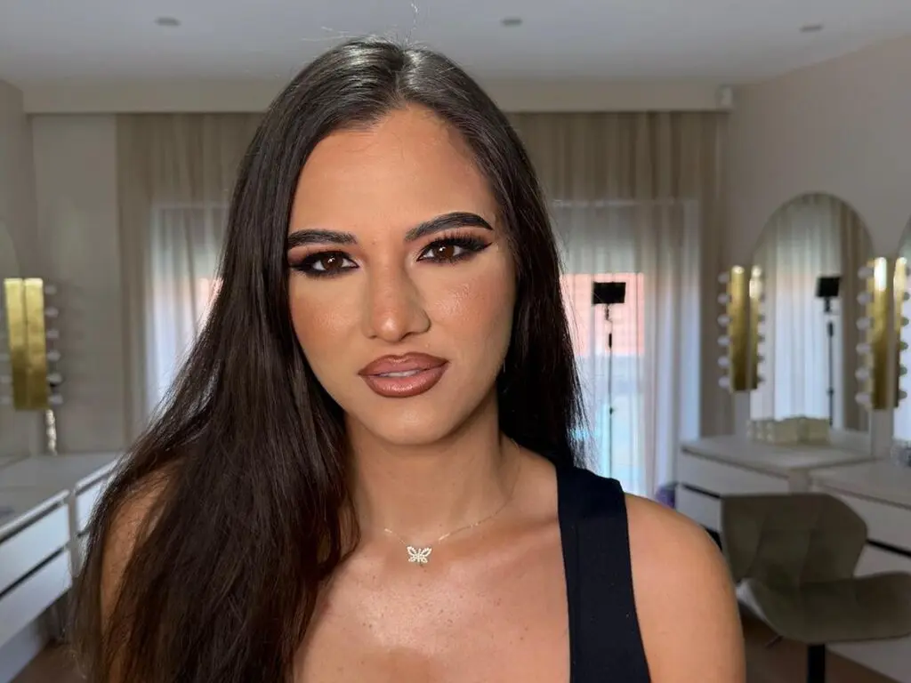 Brunette woman with glamorous smoky eye makeup wearing a black tank top in a bright, modern room.