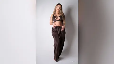 Fashionable woman in a brown crop top and wide-leg trousers posing against a minimalist studio background.