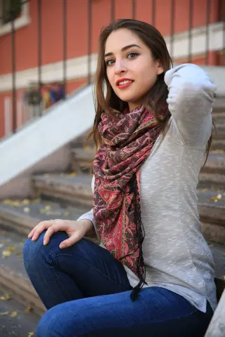 Smiling young woman in a white long-sleeve shirt and patterned red scarf sitting on outdoor stone stairs.