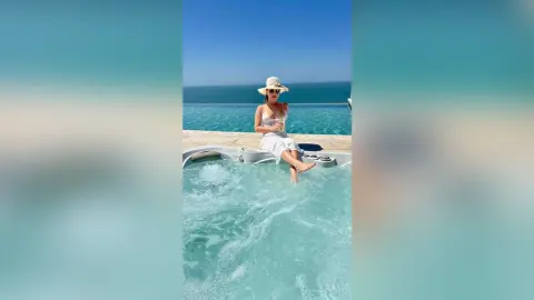 Relaxed woman in a white summer dress and sun hat relaxing in an infinity pool overlooking the blue ocean.