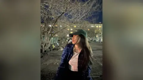 Young woman in a blue plaid shirt and green cap sitting outdoors near trees at night.