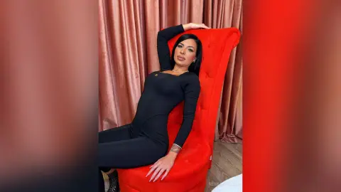 ReynaMorel poses in a tight black bodysuit while sitting on a tall red chair against pink curtains.