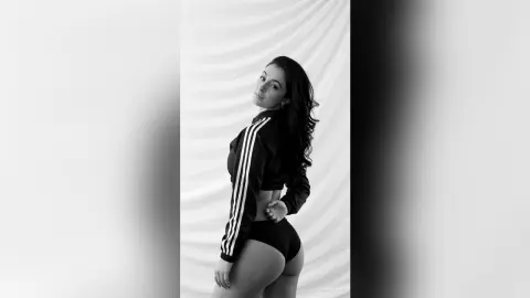 Glamorous woman in a black Adidas tracksuit posing against a white draped backdrop in a moody black and white portrait.