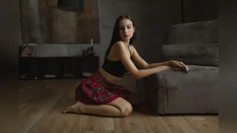 Young woman in a black crop top and red plaid skirt sitting on a floor in a moody, dimly lit room.