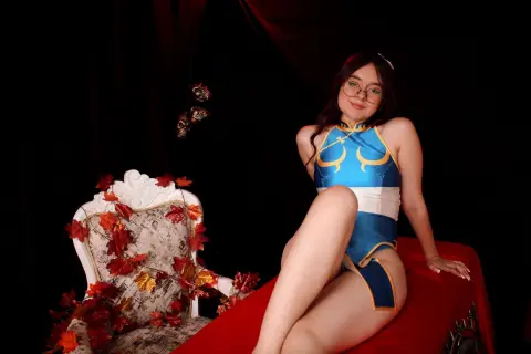 Smiling woman in blue and gold cosplay sitting on a red velvet lounge surrounded by autumn leaves.