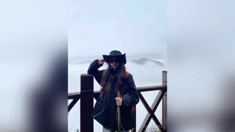 Woman in black jacket and hat standing on a wooden balcony overlooking a misty mountain landscape.
