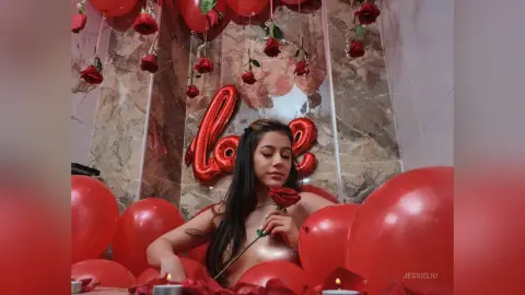 JessieLiu poses naked behind a red rose, surrounded by red balloons and red roses against a marble wall.