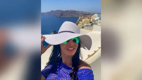 MissyJolie smiles wearing a wide-brimmed white sun hat and sunglasses, dressed in a blue lace top against a coastal Santorini background.
