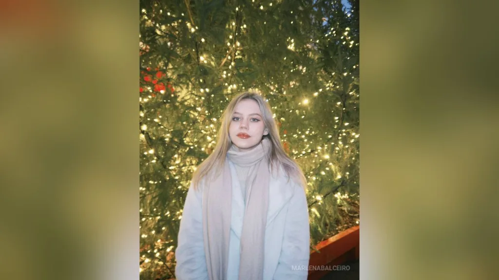 MarlenaBalceiro smiles with her long blonde hair and light eyes, wearing a black top in a bright room with a green plant behind her.