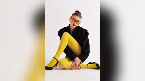 Fashionable woman in black blazer and bright yellow tights posing confidently against a white studio background.