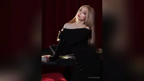Elegant blonde woman in a chic black off-the-shoulder dress posing against a moody red curtain background.