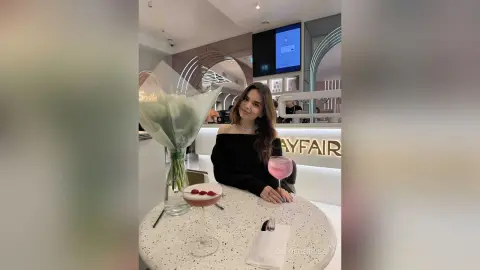Young woman in a black off-the-shoulder top enjoying a pink cocktail at a modern, bright cafe.