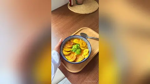 A person holding a blue bowl of golden vegetable soup topped with fresh herbs on a wooden tray.