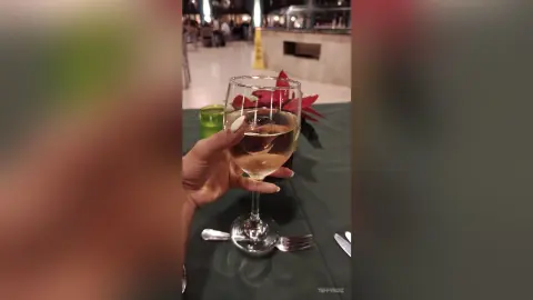 Hand holding a glass of white wine at a dimly lit restaurant table with festive red flowers.