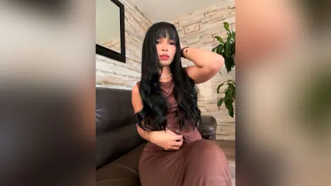 Young woman with long dark hair in a brown dress sitting on a leather sofa in a modern living room.