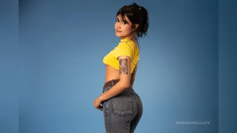 Young woman in a yellow crop top and denim jeans posing against a blue studio background.