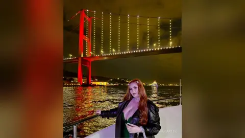 Redheaded woman in a black leather jacket on a boat at night with a glowing suspension bridge background.