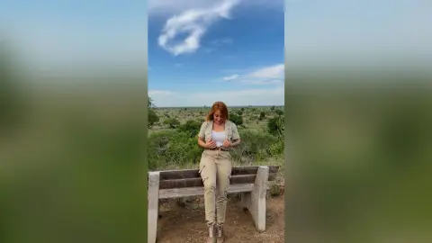 Redheaded woman in beige cargo pants and white top sitting on a park bench under a blue sky.