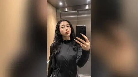 Young woman with long wavy hair in a black zip-up jacket taking a moody mirror selfie in an elevator.