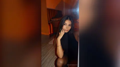 SamanthaBrazer poses in a black top with her hand touching her face, showing her long dark hair in a dimly lit room.