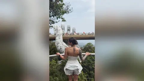 EmiliaRhodes stands on a balcony overlooking a giant stone hand statue in a lush green forest.