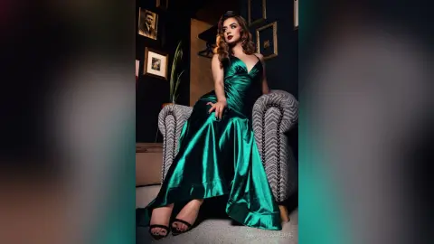 Elegant woman in a shimmering emerald green satin gown sitting on a vintage armchair in a moody, dark room.