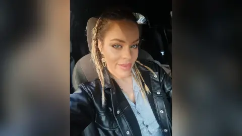 Smiling woman with blonde braids wearing a black leather jacket sitting in a car interior.