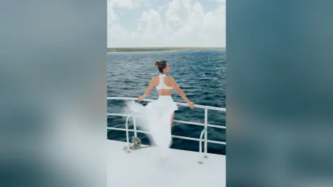 Elegant woman in a white flowing dress standing on a boat deck overlooking the blue ocean under a sunny sky.